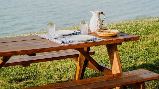 A wooden picnic table with a pitcher, glasses, and a plate set on a grassy area by a body of water. - Waterfront Rappahannock River Cottage, Private Pool, Kayaks, Fire Pit & Garden (Locust Hill)