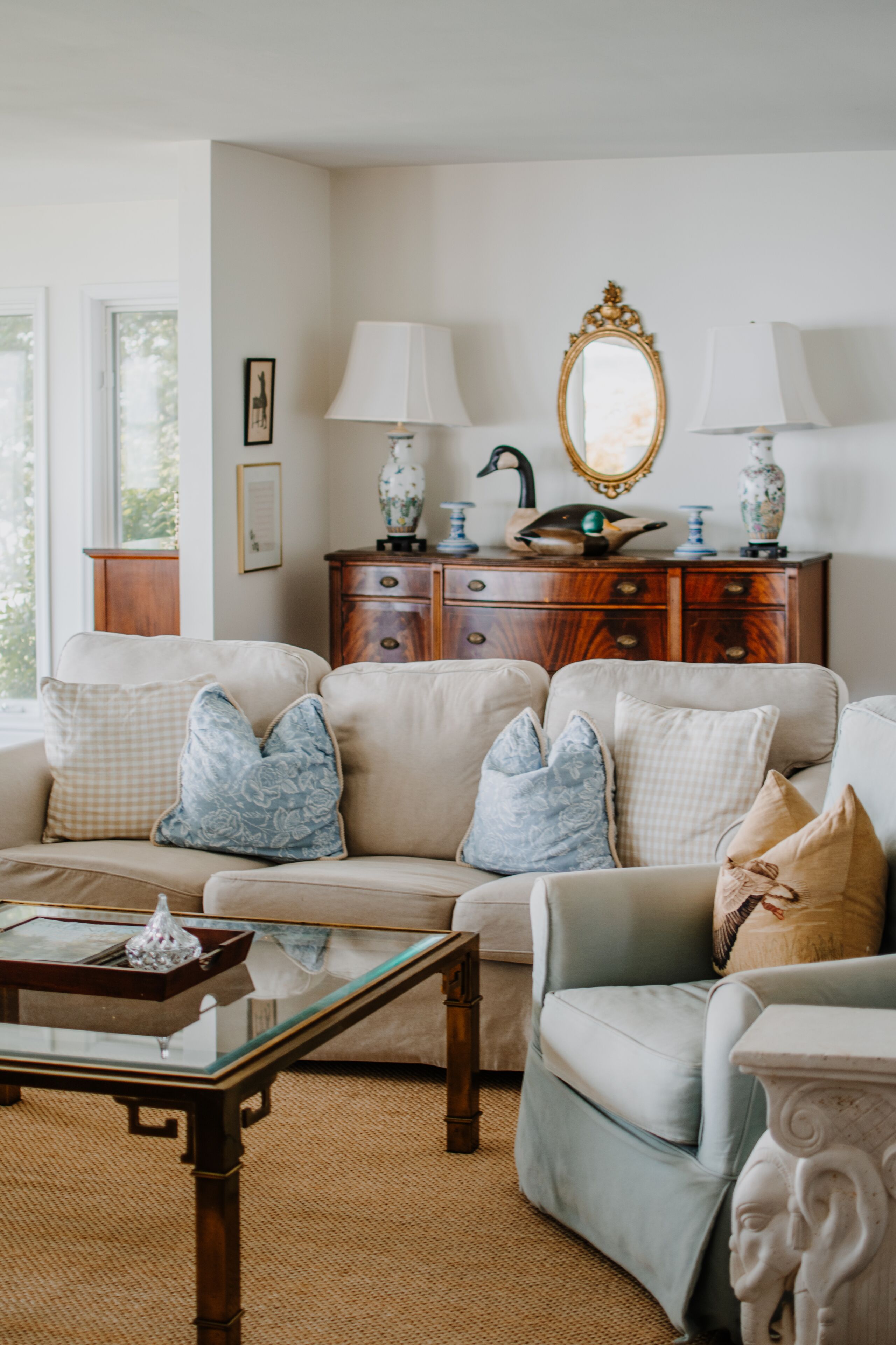Un salon avec un canapé beige, une table basse en verre et un meuble en bois orné d’objets décoratifs.