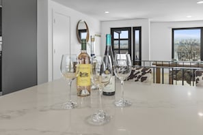 Una mesa con cubierta de mármol, botellas y copas de vino, un espejo y vistas del exterior a través de una ventana.