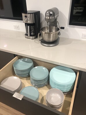 Un plan de travail de cuisine avec une machine à café, un robot pâtissier et un tiroir rempli d’assiettes empilées.