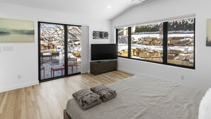 Una camera da letto moderna con un letto grande, una televisione a schermo piatto e una porta scorrevole in vetro che dà su uno spazio esterno con vista su rocce e alberi innevati.