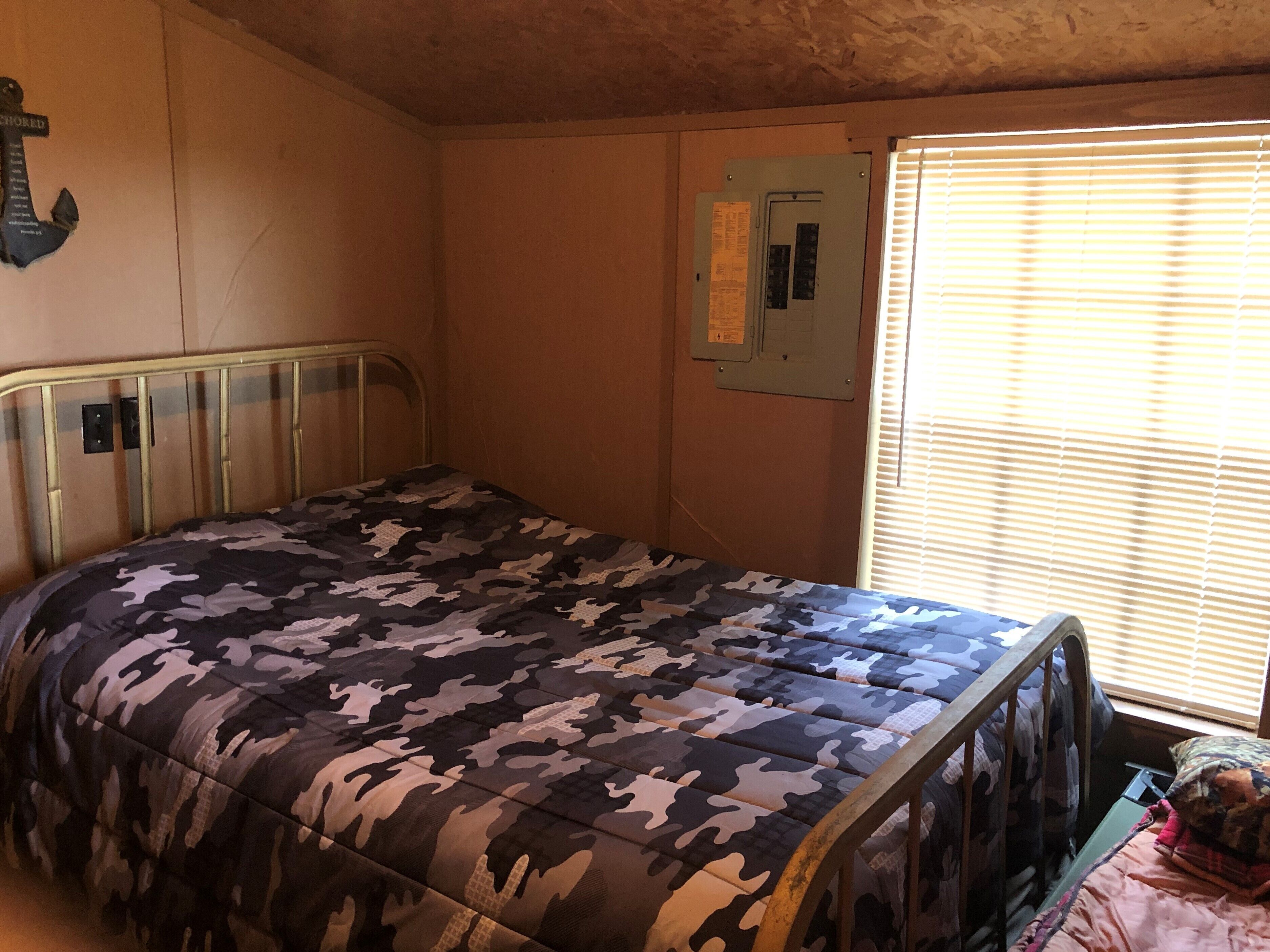 A bedroom with a bed, a window with blinds, a wall-mounted electrical box, and a cross on the wall.