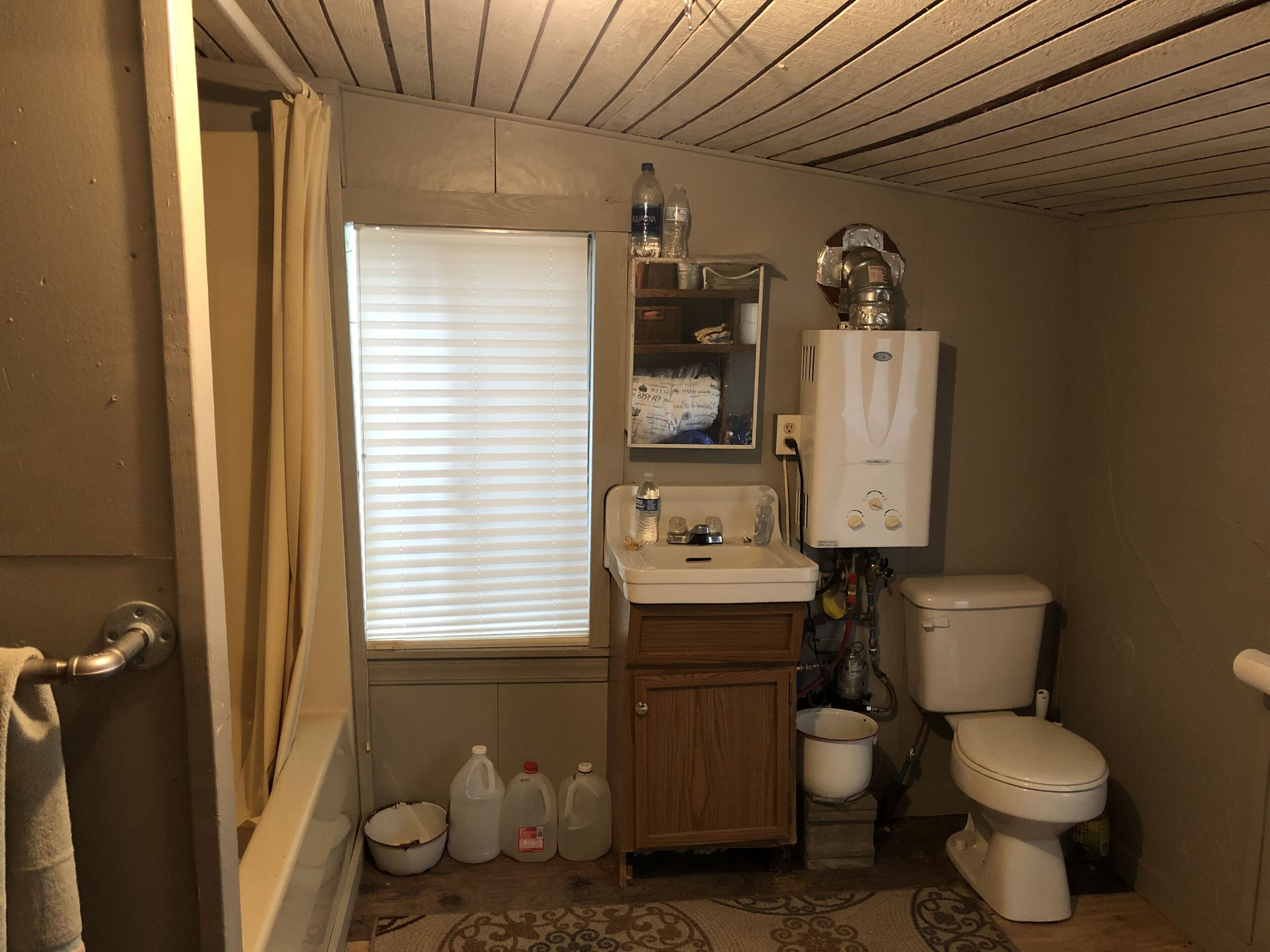 A small bathroom with a toilet, sink, and window with blinds. There are several bottles and a water heater on the wall.