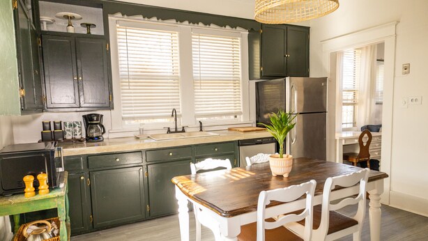 A kitchen with dark green cabinets, a wooden dining table with white chairs, a stainless steel refrigerator, and a hanging light fixture. - J Birds' Bungalow (Memphis)