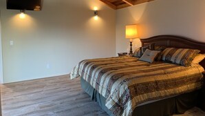 A bedroom with a wooden ceiling, a bed with a patterned bedspread, a nightstand with a lamp, and a wall-mounted TV. - Taylor II House, ocean view, close to everything (Crescent City)