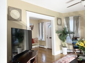 A living room with a flat-screen TV on a wooden stand, a potted plant, a chair with a striped cushion, and a patterned rug. - Beautiful Retreat close to Downtown Boston. Pets friendly. (Boston)