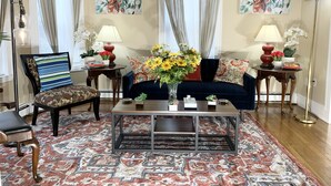 Sala de estar com um sofá azul escuro, poltronas estampadas, uma mesa de centro e um tapete padronizado. Há duas luminárias, um vaso com flores e um quadro na parede.