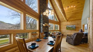 A wooden cabin with a dining area featuring a large table and chairs, a stone fireplace, and large windows offering views of the surrounding landscape. - Custom Log Cabin Close to Cades Cove in GSMNP! (Townsend)