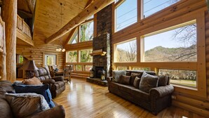 A log cabin interior with a stone fireplace, wooden furniture, and large windows offering a view of the outdoors. - Custom Log Cabin Close to Cades Cove in GSMNP! (Townsend)