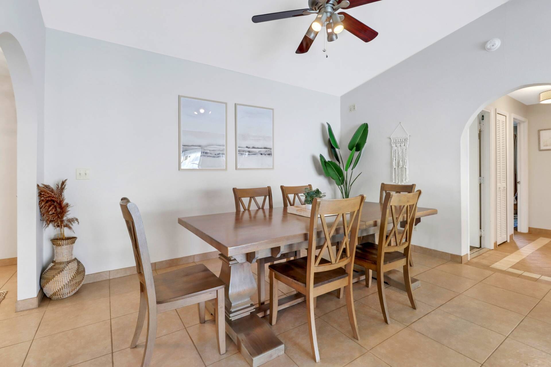 Un coin repas avec une table et des chaises en bois, un ventilateur de plafond et deux tableaux encadrés au mur.