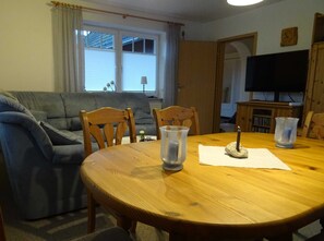 A living room with a wooden dining table, two chairs, a grey sofa, a TV, and a window with blinds.