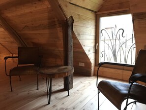 A cozy attic room with wooden walls, a sloped ceiling, and a large window with decorative iron bars. There are two chairs and a small round table.