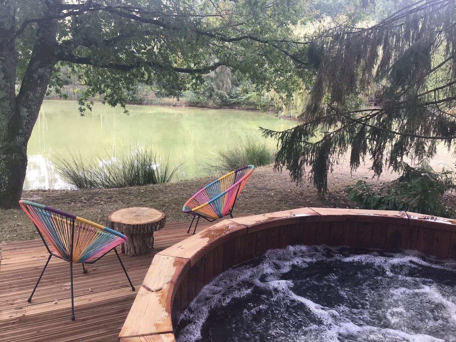 A hot tub with two colorful chairs on a wooden deck, overlooking a calm pond.