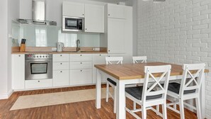 A modern kitchen with white cabinetry, a built-in oven, microwave, and gas stove. There is a wooden dining table with white chairs. - Grand Apartments - Willa Morska (Sopot)