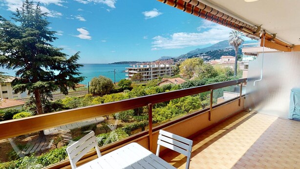 A balcony with a table and chairs, overlooking a coastal view with buildings and greenery. - Spacious studio in Menton with large terrace and garage (Menton)