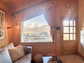 A wooden cabin interior with a sofa, a table, a window with a view of mountains, and a door. - 4-room duplex with parking and wifi at the foot of the slopes (Combloux)