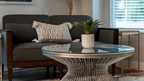Ein modernes Wohnzimmer mit einem dunklen Sofa, einem Glastisch mit außergewöhnlicher Metallkonstruktion und einer Topfpflanze.