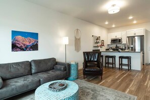 Ein modernes Wohnzimmer mit einem grauen Sofa, einem blauen Couchtisch mit Muster und einem Ledersessel. Es gibt eine Küchenzeile mit weißen Schränken und Edelstahlgeräten.