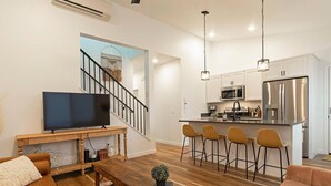 Un salon moderne avec un téléviseur, une table basse en bois et un canapé. On peut apercevoir une cuisine équipée d’un bar et d’appareils électroménagers en acier inoxydable.