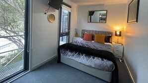 Ein Schlafzimmer mit Bett, Nachttisch, Lampe und einem Fenster mit Blick auf Bäume.
