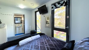 Ein Schlafzimmer mit Bett, Fernseher, einem Fenster mit Blick auf Bäume, einer Tür und einem kleinen Badezimmer mit Waschbecken und Spiegel.
