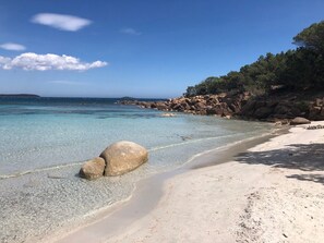 Una playa con agua cristalina, rocas grandes y un acantilado rocoso con árboles.
