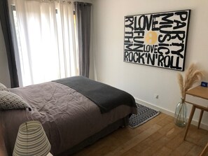 Une chambre à coucher avec un lit, une œuvre d’art murale, une petite table avec un vase et une fenêtre avec des rideaux.