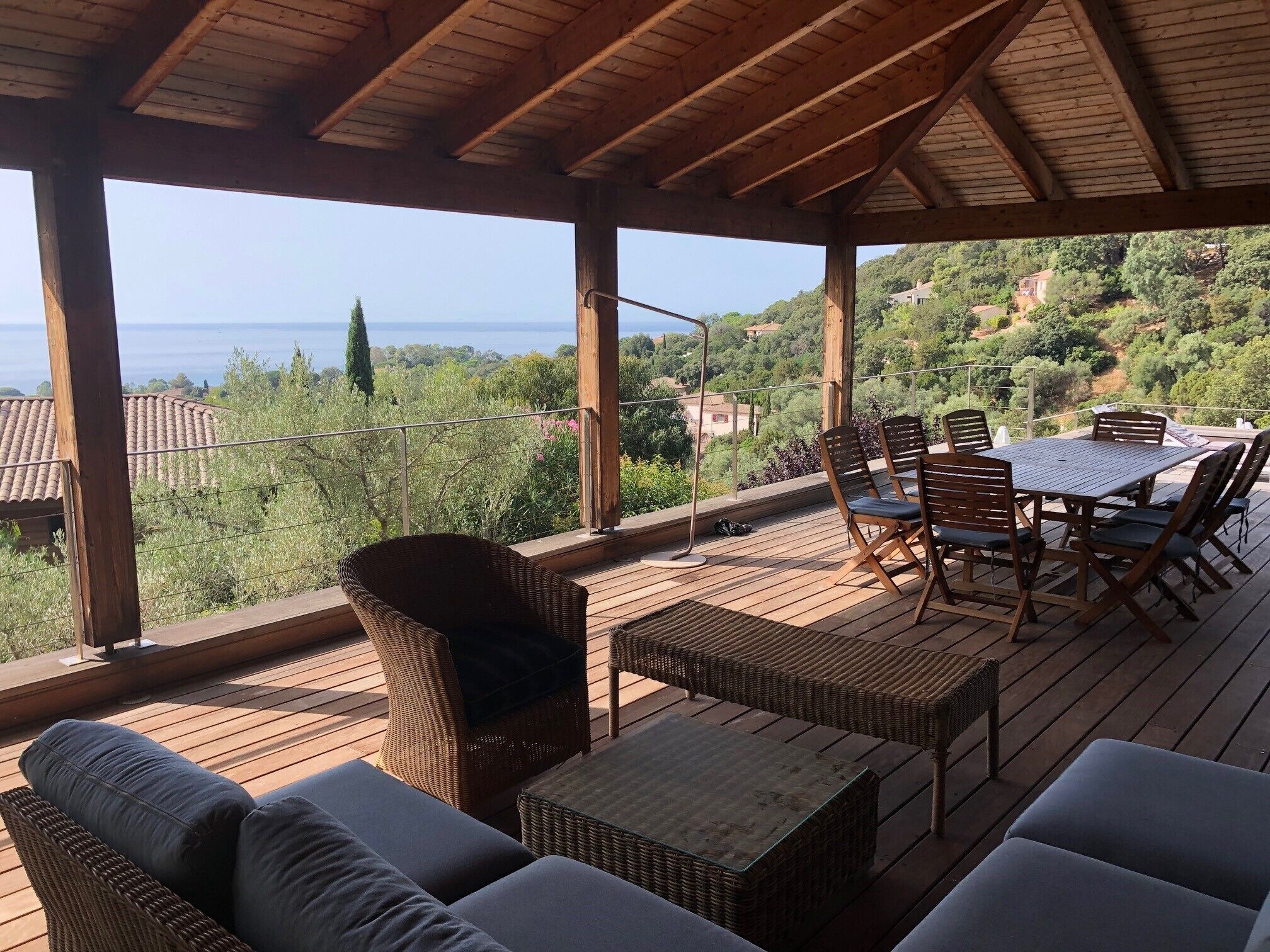 A covered outdoor seating area with wicker furniture and a table, overlooking a scenic landscape with hills and a body of water.