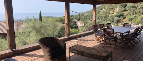 Un área de estar al aire libre cubierta con mobiliario de mimbre y una mesa, con vistas a un paisaje pintoresco con colinas y una masa de agua.
