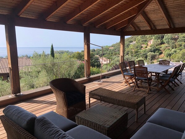 Un espace extérieur aménagé, abrité et doté de meubles en osier et d’une table, offrant une vue panoramique sur un paysage pittoresque avec des collines et une étendue d’eau.