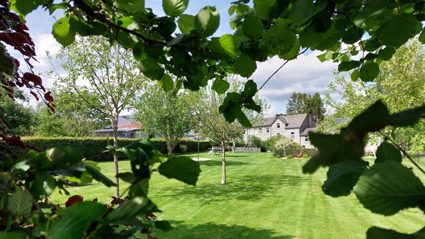 A well-maintained grassy lawn with a hedge and trees in the foreground, leading to a stone building with a traditional design.