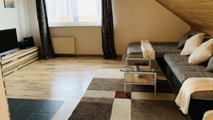 Un dormitorio acogedor en el ático con techo inclinado, un ventanal con cortinas, un sofá con cojines, una mesa de centro de vidrio y un televisor sobre una estantería de madera.
