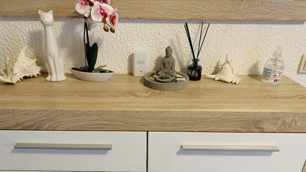 Una encimera de cocina con una figurita de gato blanco, una orquídea en maceta, una estatua de Buda, un difusor y una botella blanca.