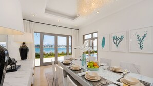 A modern dining area with a glass table, white chairs, and a view of the sea. - Maison Privee - Frond Villa K (Dubai)