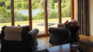 Una habitación con un amplio ventanal con vistas a un lago, dos sillones, una mesa de madera y un jarrón con flores.