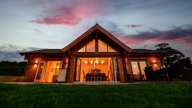 A wooden cabin with a large open front door, a dining area inside, and a view of the sky with pink clouds. - Cuddfan Lodge on a Beautiful Lake (Newport)