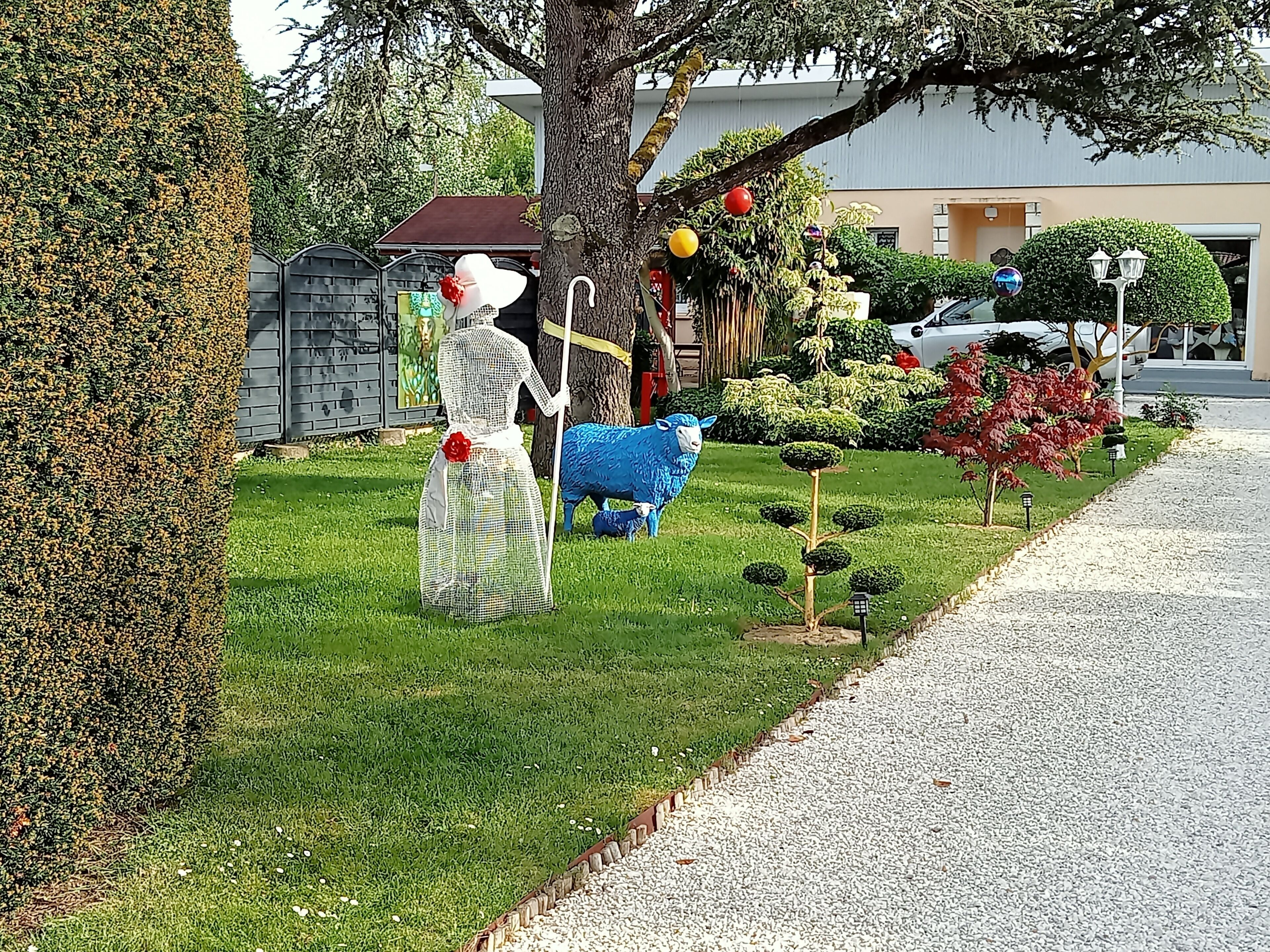 A garden with a statue of a person holding a staff, a blue sheep sculpture, and a small tree with spherical topiary.