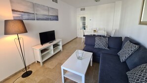 Een woonkamer met een televisie op een witte dressoir, een donkerblauwe bank met gestreepte kussens, een witte salontafel en een vloerlamp.