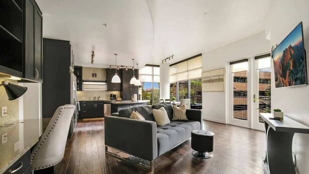 A modern living room with a dark grey sofa, a white armchair, and a black ottoman. There is a kitchen area with dark cabinets and pendant lighting. Large windows provide natural light and a view of the outdoors. - Walkable Uptown Dallas 3BR with Pool, Pets, Wi-Fi (Dallas)