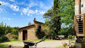 A rustic stone building with a small door and a chimney, surrounded by greenery and a wheelbarrow on a gravel path.