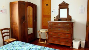 A room with a wooden wardrobe, a dresser with drawers, a small table, and a bed with a patterned bedspread.