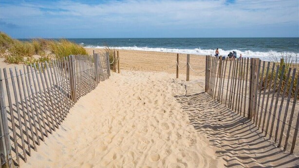 Una spiaggia sabbiosa con un recinto di legno che conduce all'acqua, e persone che camminano lungo la riva.