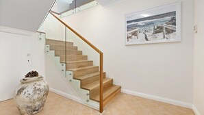 Eine Treppe mit Glastrennwand und Holzboden, eine weiße Vase mit Trockenblumen und ein gerahmtes Gemälde einer Küstenlandschaft an der Wand.