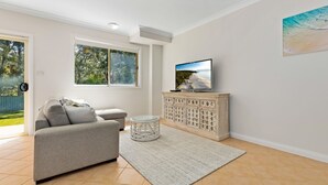 Ein Wohnzimmer mit grauem Sofa, einem Glastisch, einem Schrank mit dekorativen Verzierungen, einem Flachbildfernseher auf einem Standfuß und einem großen Fenster mit Blick auf Bäume und einen Zaun.
