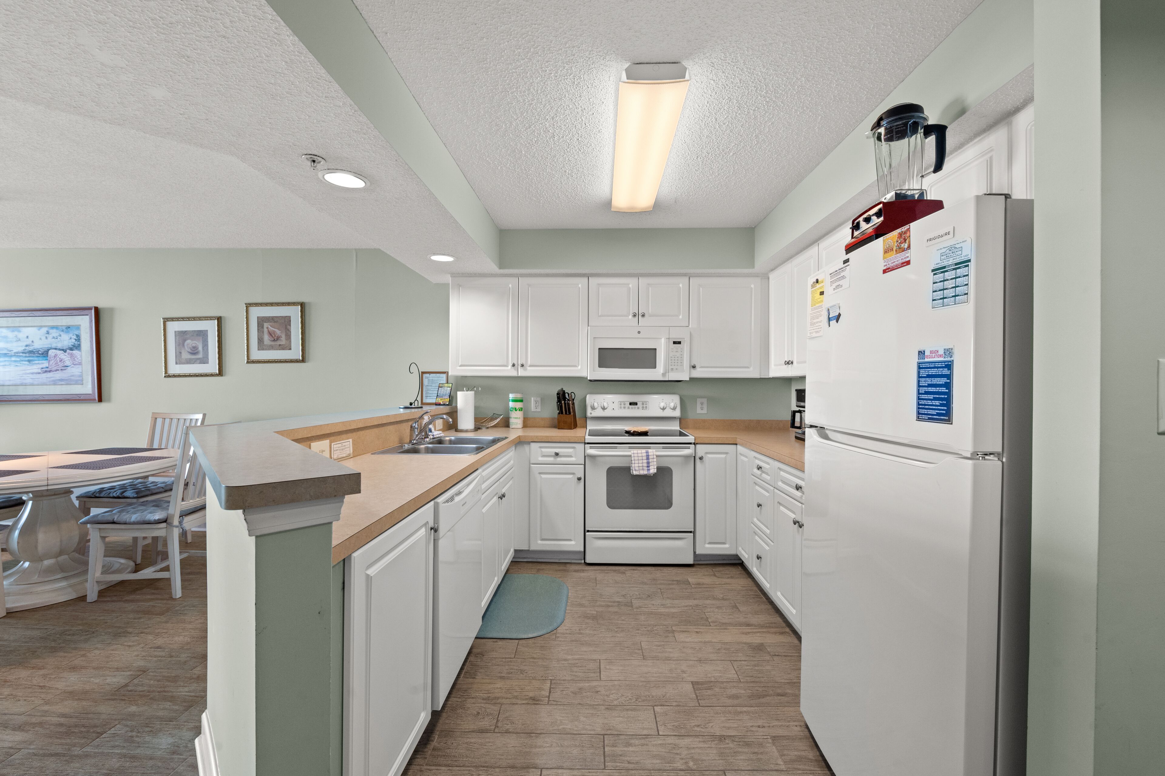 A modern kitchen with white cabinets, a built-in oven, microwave, and refrigerator. There is a dining area with a table and chairs, and a blue rug on the floor.