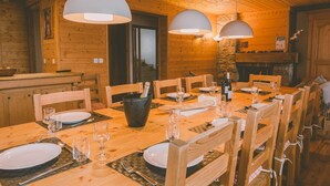 A wooden dining room with a large table set for six, surrounded by wooden chairs. There are two pendant lights hanging above the table, and a stone fireplace is visible in the background.