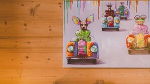 A wooden cabin-style room with a bed, pillows, a nightstand, and a painting of colorful vehicles on the wall.