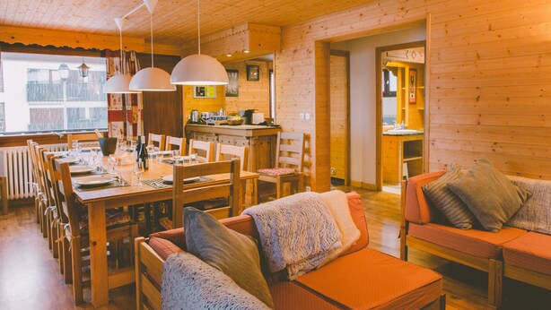 A cozy wooden interior with a dining area, a sofa, and a kitchen visible through an open doorway.