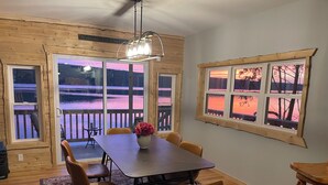 A cozy dining area with a wooden table, chairs, and a vase of flowers. Large windows offer views of a sunset over water.
