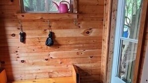 A wooden cabin interior with a bench, a small window with a pink kettle, and a door leading outside.